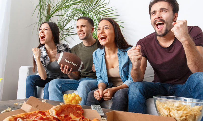 Picture of two women and two men smiling and cheering with a football in one person's hand and food on the table in front of them indicating that are cheering for American football