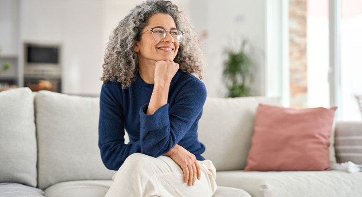 Woman with curly gray hair in dark blue sweater sits on couch with chin resting on fist as she looks of camera