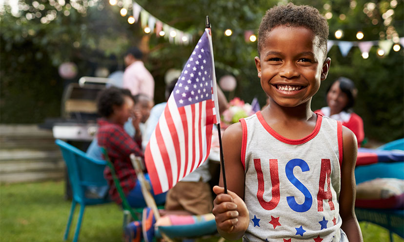 Kid smiling while holding an American flag and wearing a USA shirt. Family at BBQ and table in background